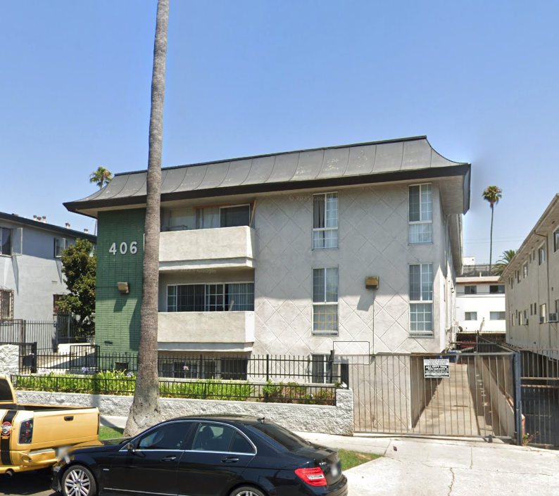 A black car is parked in front of a two-story building with a green door and a palm tree in front of it.